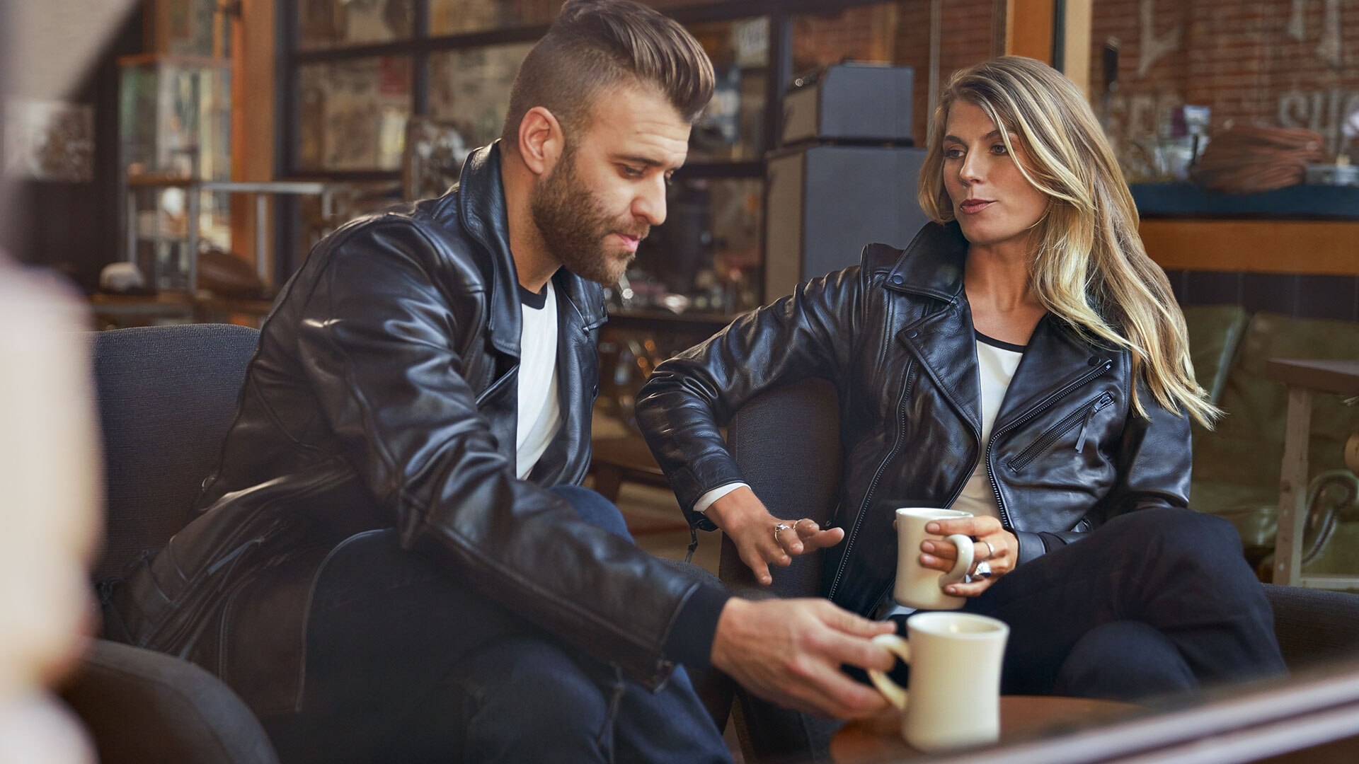 Auf dem Bild sitzen sich ein junger Mann in der Lederjacke FlatTwin für Herren und eine junge Frau in der Lederjacke FlatTwin für Damen gegenüber.