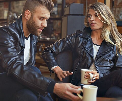 Auf dem Bild sitzen sich ein junger Mann in der Lederjacke FlatTwin für Herren und eine junge Frau in der Lederjacke FlatTwin für Damen gegenüber.