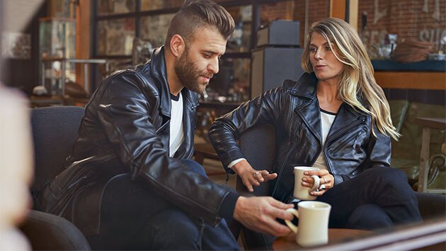 Auf dem Bild sitzen sich ein junger Mann in der Lederjacke FlatTwin für Herren und eine junge Frau in der Lederjacke FlatTwin für Damen gegenüber.